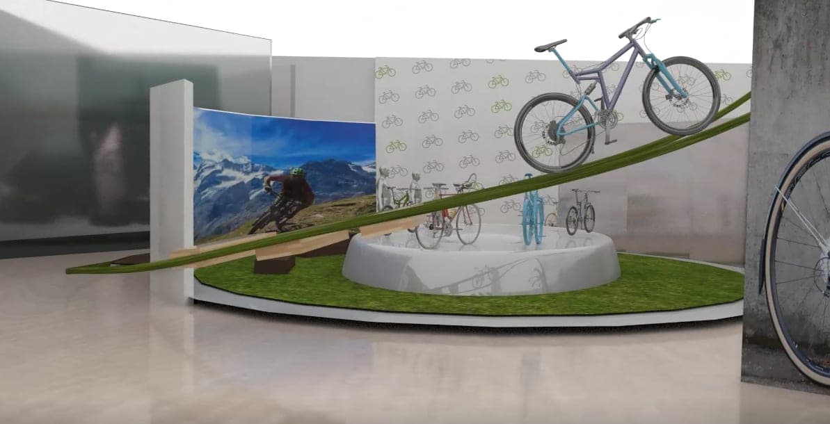 3D render of the central GOGOBIKE display: bikes on a round white platform with a grass ring, a green ramp carrying a mountain bike, and an alpine backdrop.