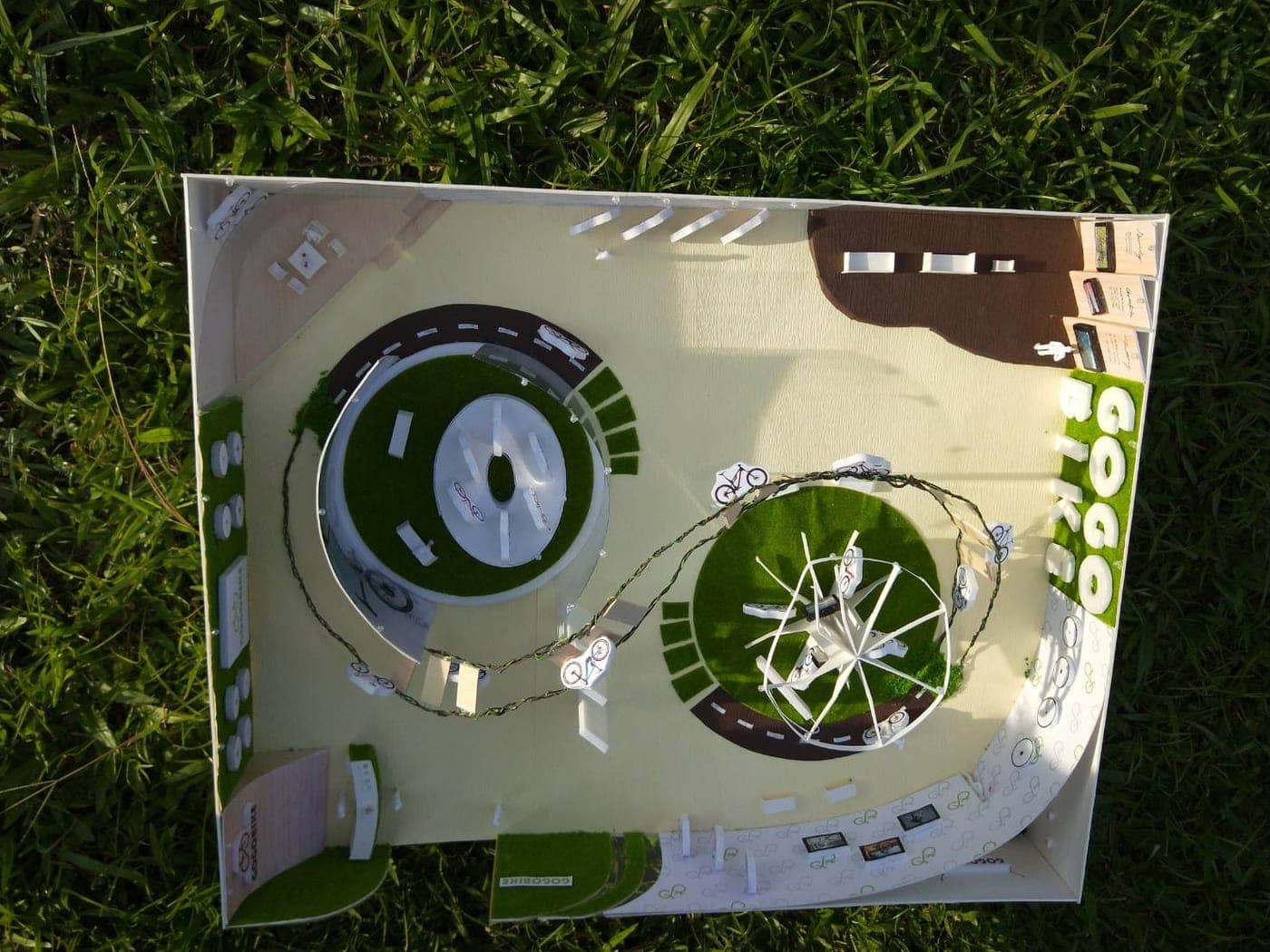 Overhead photo of the handmade GOGOBIKE exhibition scale model on grass, with two circular display zones, turf details, and GOGO BIKE lettering