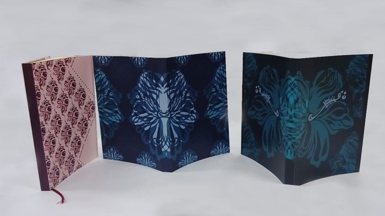 Three handmade leaf-pattern notebooks standing upright with covers fanned open, in pink, navy blue, and dark teal printed designs.