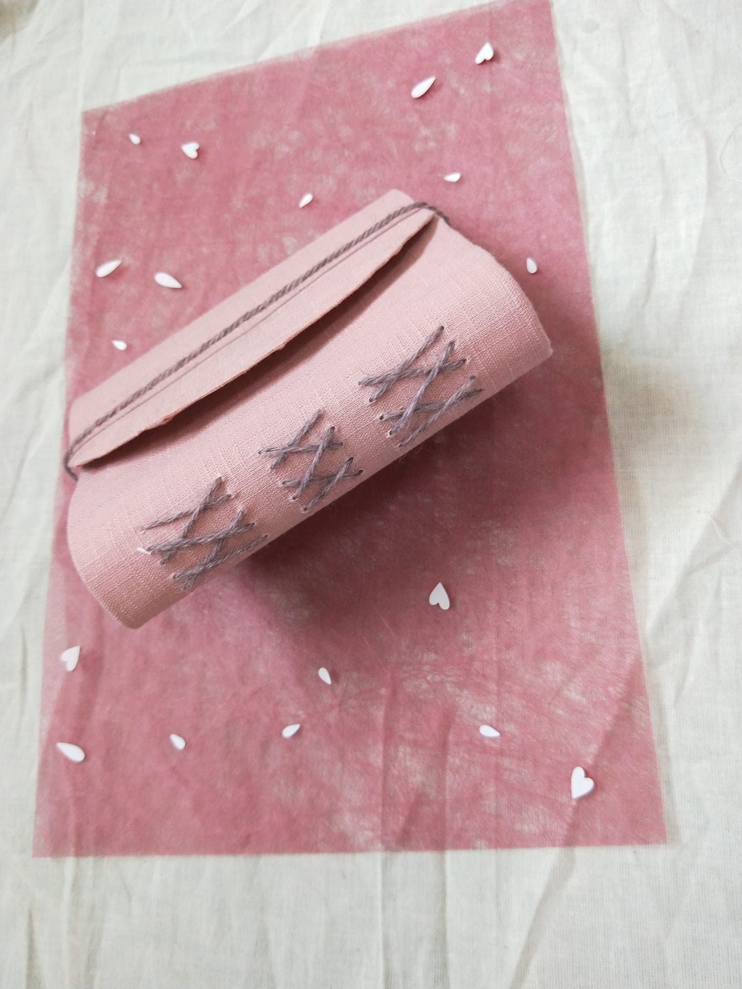 Pink linen handbound book with grey cross-stitch binding along the rounded spine and a yarn tie, on pink paper scattered with white paper hearts.