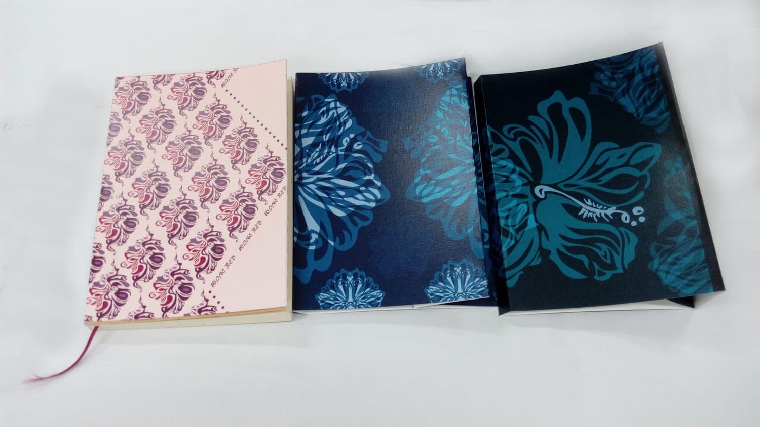 Top-down view of three handmade notebooks with printed leaf-pattern covers in pink, navy, and dark teal; one has a red ribbon bookmark.