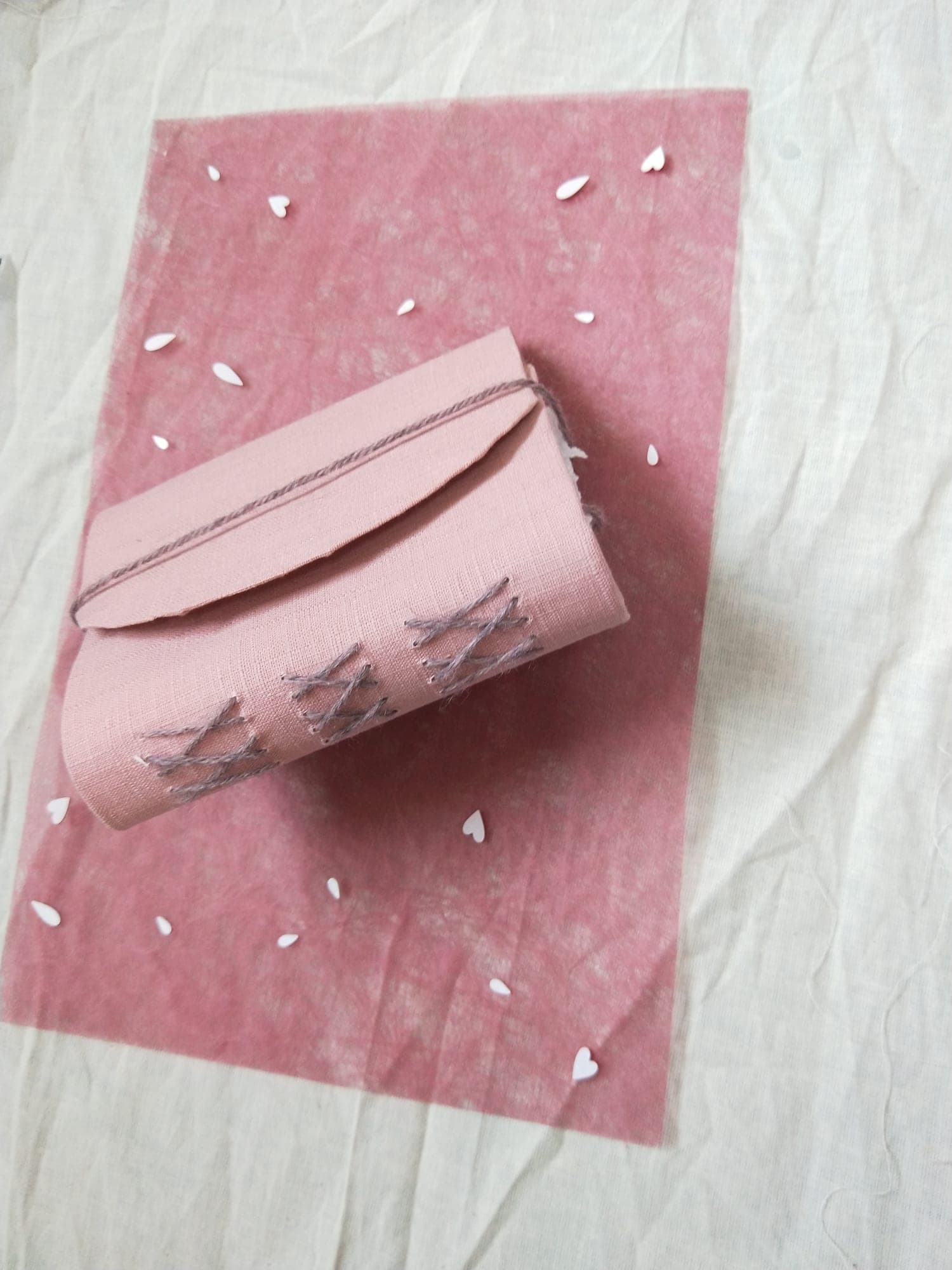 Pink linen handbound book with grey cross-stitch spine binding and yarn tie, shown slightly wider on pink paper with white hearts.