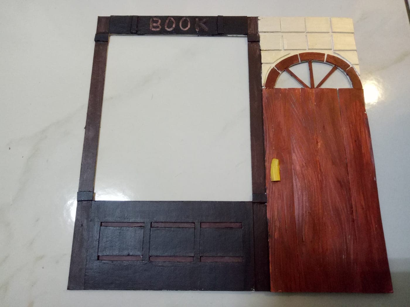 Handmade miniature bookshop facade: dark timber frame with hand-lettered BOOK sign, plank-textured red door with yellow handle, fan transom, and cream brick wall.