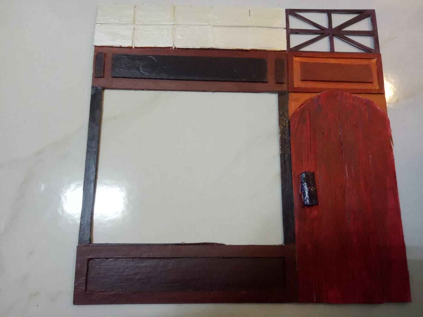 Handmade miniature storefront facade in painted cardboard: arched red wood-grain door, dark signboard, large window opening, and white shingle trim along the top.
