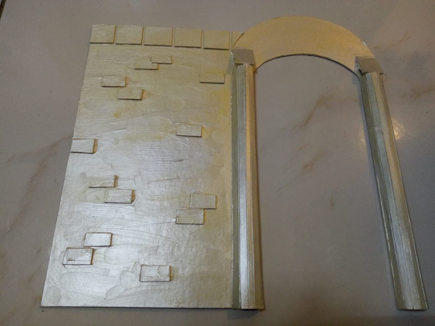 Work-in-progress model parts: a pale brick-relief wall panel beside an unpainted arched doorway frame with slender columns, laid out on a tabletop.
