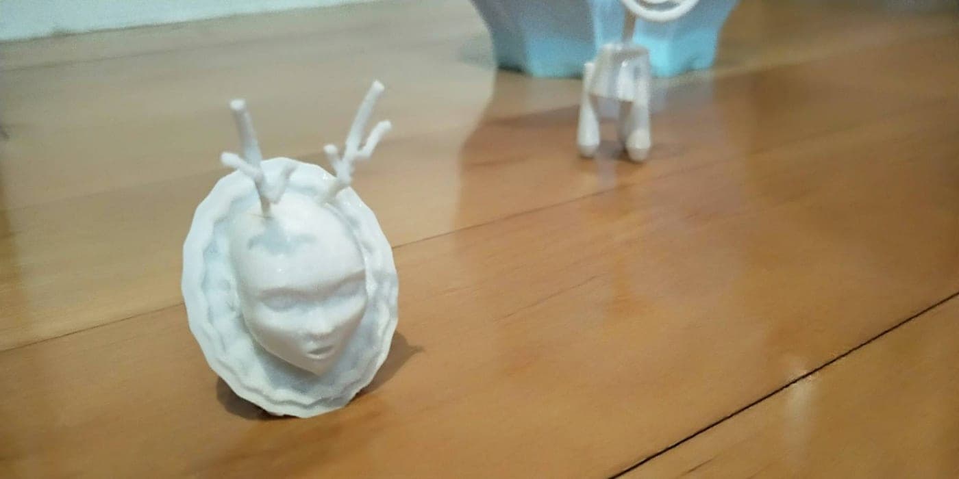 White 3D-printed face relief plaque with antler-like supports in the foreground; a blue low-poly vase and a small four-legged print stand blurred behind it.