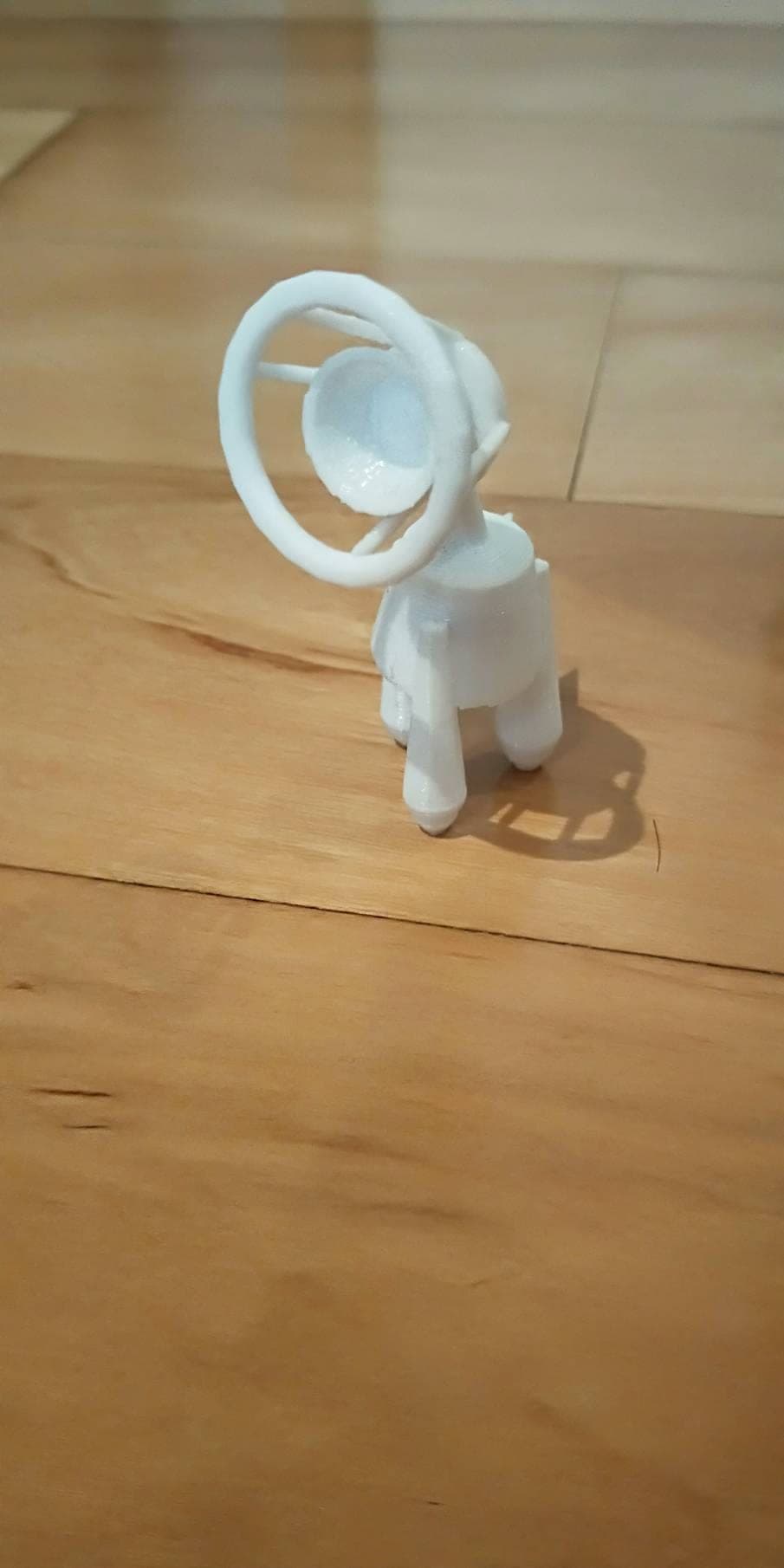 White 3D-printed four-legged figure with a round dish head encircled by a ring, standing upright on a wooden floor in warm light.