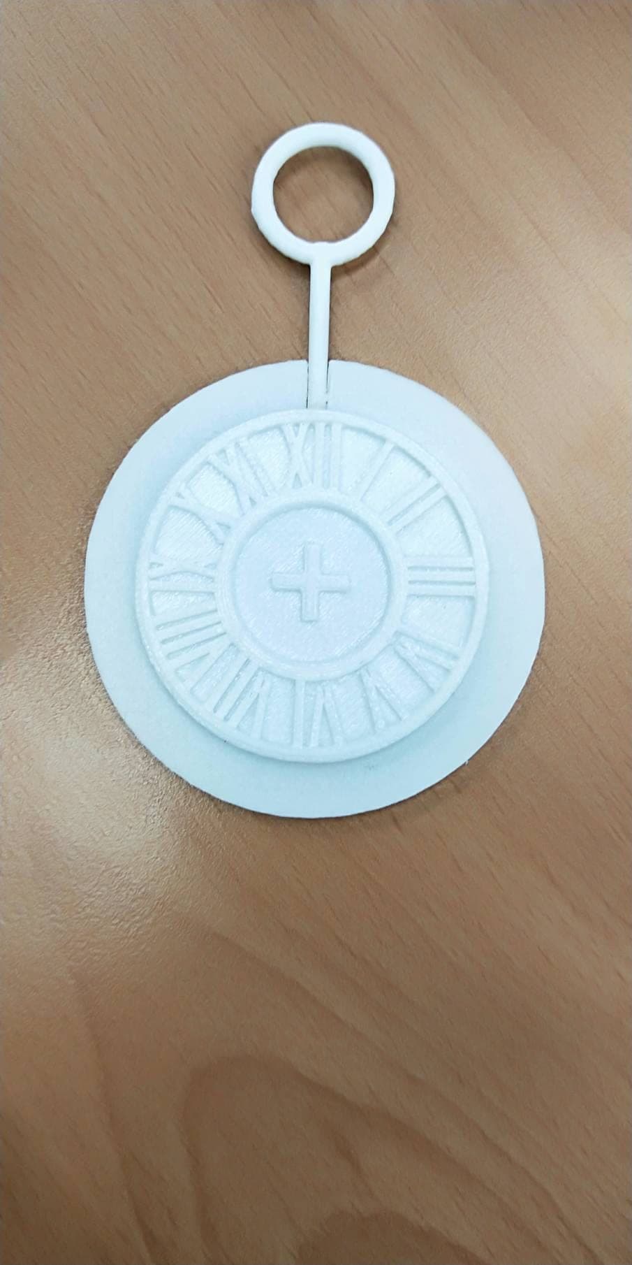 White 3D-printed pocket watch charm with a Roman numeral dial and a raised cross at its center, with a hanging ring, on a wooden desk.